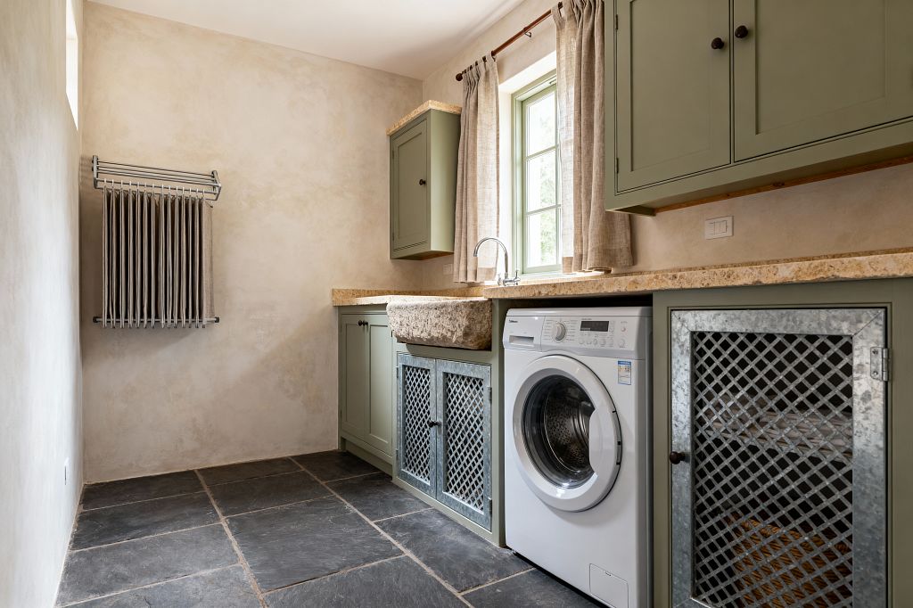 A farmhouse-style laundry room with sage green cabinets and a beige limestone countertop, centered by a rough-hewn stone trough sink and a white front-load washing machine. The space has limewashed plaster walls, a window with green trim and natural linen curtains, dark charcoal slate floor tiles, galvanized lattice cabinet doors for ventilation, and a wall-mounted metal drying rack, all lit by soft natural daylight.
