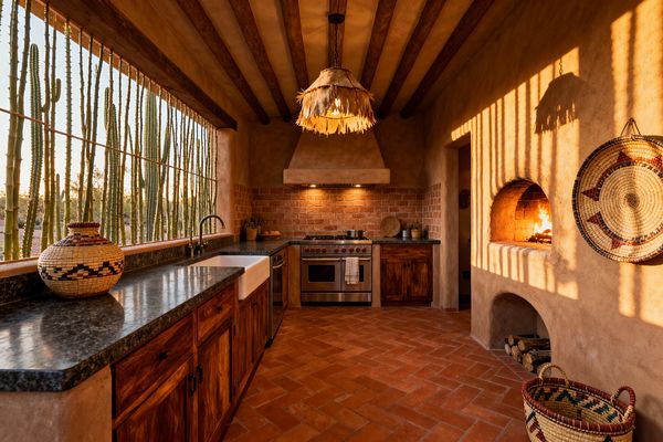 A Southwestern kitchen with plaster adobe walls, exposed wood ceiling beams, and a terracotta herringbone floor. Dark stone countertops top rustic wood cabinets beside a white farmhouse sink, with a red brick backsplash and plaster range hood framing stainless appliances. A domed kiva-style fireplace glows on the right, woven baskets decorate the walls, and a fringe pendant hangs overhead. A wide window lined with vertical cacti floods the space with warm desert light.
