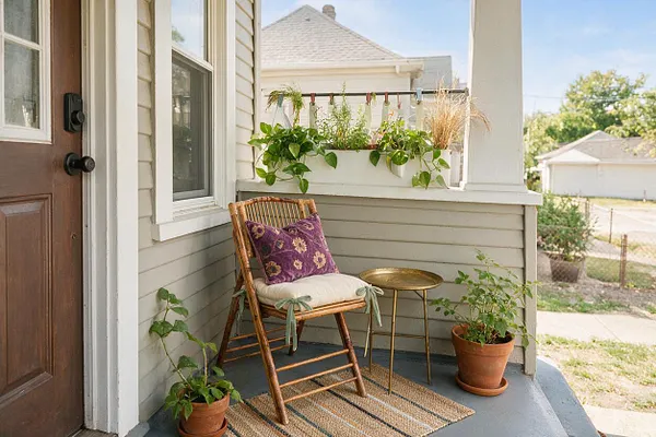 A small front porch with gray flooring and light beige siding features a wicker chair with purple patterned and cream cushions, a brass side table, and several terracotta potted plants. A woven natural rug anchors the seating area, while a wooden front door and hanging planters add warmth and a casual bohemian feel.