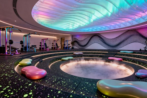 A psychedelic-inspired home gym features a circular sunken pit emitting mist, encircled by tiered black speckled steps lit with continuous LED strips. Above, an undulating translucent ceiling glows in cyan, pink, and purple gradients, while iridescent pebble-shaped poufs dot the seating. Along the back wall, rainbow powder-coated weight machines line up beneath white walls with a wavy mesh art installation and perimeter cove lighting.