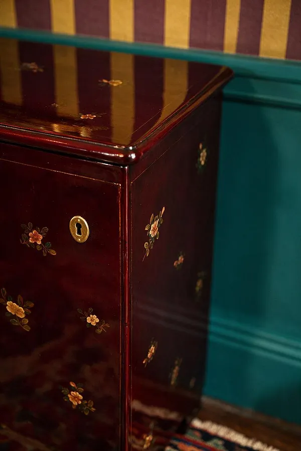 A close-up of a glossy burgundy lacquer cabinet decorated with tiny painted floral motifs and a brass keyhole plate. It sits against a teal wall with gold-and-plum vertical striped wallpaper, creating a rich maximalist contrast.