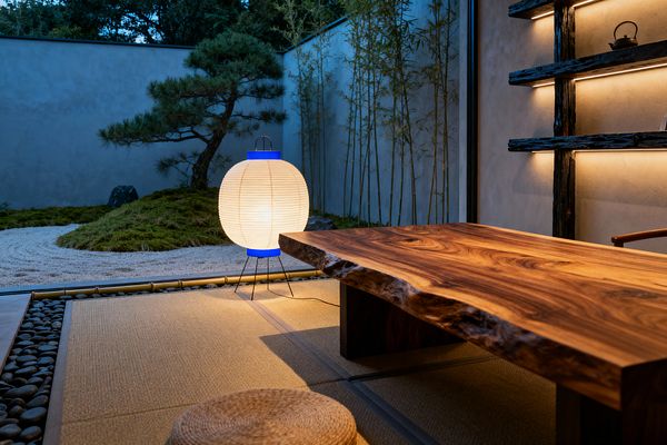 A serene Japanese-style home office features a low live-edge wooden desk set on tatami mats beside a floor-to-ceiling window. A round washi paper lantern with blue accents glows near a pebble-and-bamboo threshold, while outside a minimalist courtyard shows raked gravel, moss mounds, a sculpted pine, and slender bamboo; inside, textured plaster walls and dark timber shelves with integrated LED lighting create a calm, balanced workspace with a woven floor cushion and teapot detail.