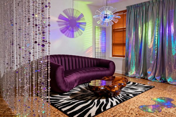 A psychedelic living room features a deep purple velvet, channel-tufted sofa on a black-and-white zebra rug. A glossy, organic burl-wood coffee table sits center stage. Iridescent metallic drapes and a beaded curtain sparkle as a clear acrylic disc pendant projects rainbow shadows on white walls. Warm wood blinds and cork flooring ground the space, while a small puddle-shaped holographic accent decorates the floor near the window.