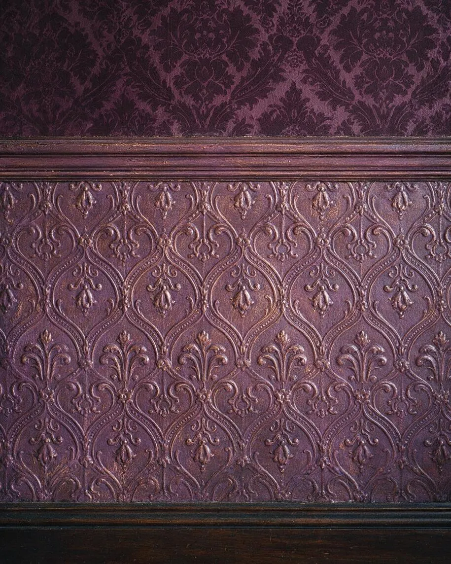A close-up of a Victorian-style wall treatment in deep plum and mauve tones. The image shows a richly embossed damask-like pattern above dark painted wood trim, creating a layered, ornate surface with a historic feel.