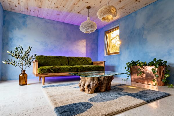 A biophilic living room features a moss‑green cushioned oak sofa set against sky‑blue limewash walls with a subtle purple glow behind it. A glass coffee table on a sculptural driftwood base sits on a cream and denim‑blue geometric rug, beneath two cloudlike white pendants and a pine tongue‑and‑groove ceiling. A copper planter with trailing vines stands near a wood‑framed window, while an amber glass bottle holds an olive branch on the floor.