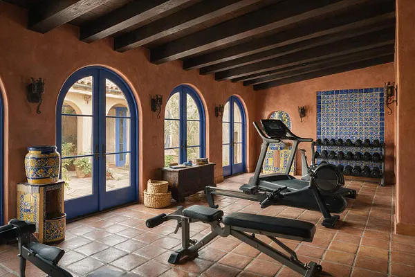 A Mediterranean-style home gym with terracotta tile floors, exposed dark wood ceiling beams, and warm stucco walls. Blue arched French doors and windows bring in natural light, while a treadmill, weight bench, dumbbell rack, and decorative blue-and-yellow tile accents add a functional yet elegant resort-like feel.