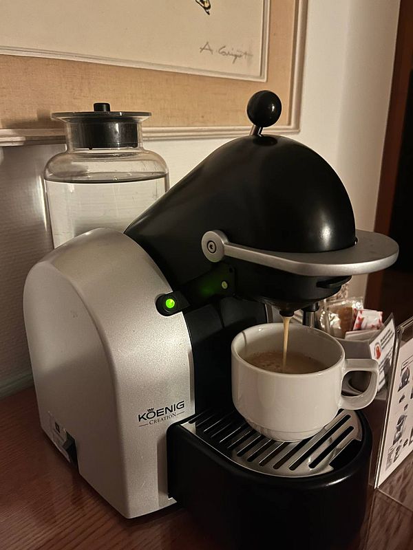 A coffee machine is brewing coffee into a white cup in a hotel room setting.