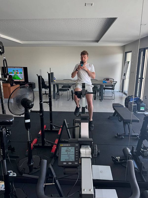 A person is seated on a rowing machine in a home gym, taking a selfie while surrounded by various gym equipment.