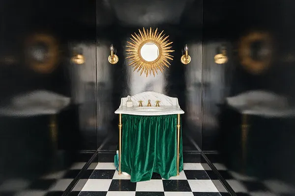 A dramatic powder room with glossy black walls, a black-and-white checkered floor, and a small marble sink wrapped in emerald green velvet. A gold sunburst mirror and matching brass sconces add Hollywood Regency glamour.