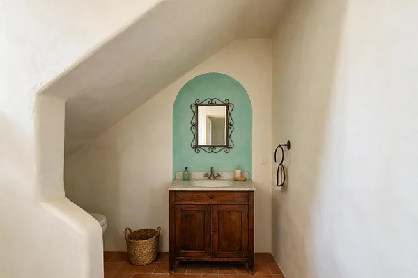 A small Mediterranean-style powder room with a sloped white plaster ceiling, terracotta tile floor, and a dark wood vanity topped with a light stone sink. An aqua arched wall niche frames a decorative mirror above the basin, while a woven basket and simple towel ring add rustic texture.