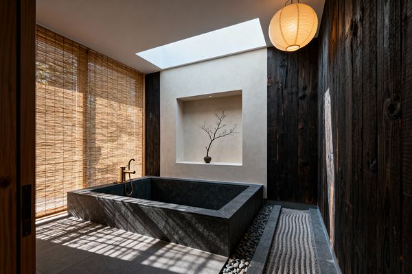 A Japanese Zen bathroom featuring a deep rectangular stone soaking tub with a brass faucet, set beside a narrow dry garden of raked gravel and river stones. Sunlight filters through bamboo blinds and a large skylight over a plaster wall niche displaying a single branch in a vase; textured charred wood clads one wall, while a paper lantern glows softly, creating a calm, minimalist atmosphere.