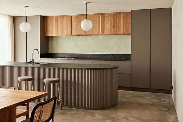A Scandinavian kitchen with a curved, fluted taupe island topped in honed dark stone with a stainless gooseneck faucet, paired with two rope-legged stools. The back wall has matte taupe base units and a tall pantry, a matching dark stone ledge with a gas hob, and natural oak upper cabinets above a pale sage limewashed backsplash. White globe pendants, an oak dining table with a cane chair, polished concrete flooring, and sheer curtains complete the calm, light-filled space.