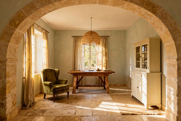 A sunlit French Country home office viewed through a limestone arch centers a rustic trestle desk under a woven rattan pendant. Pale limewashed sage walls, tumbled limestone flooring, and linen curtains frame paned windows; an olive velvet wingback sits left of the desk and a cream-painted glass-door hutch stands to the right atop a jute rug, with warm afternoon light casting patterned shadows across the room.