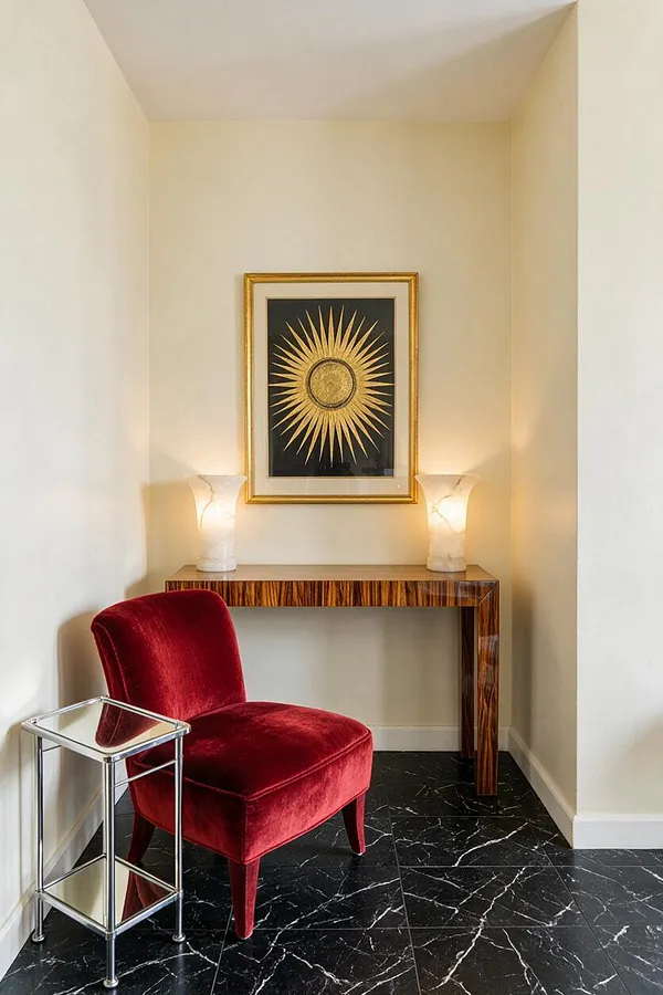 A narrow studio apartment nook styled in Art Deco fashion, featuring a red velvet accent chair on black marble-look tile flooring. A glossy wood console table sits against a cream wall, topped with two alabaster-style lamps and a framed gold sunburst artwork.