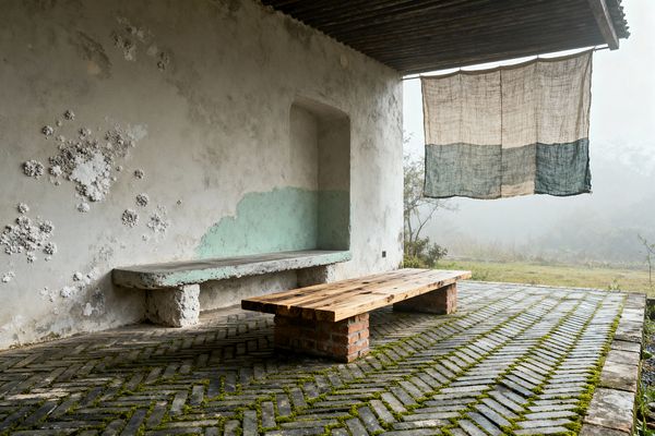 A covered patio features a weathered plaster wall in off-white with a faded mint band and crusted, textured patches, plus a built-in concrete bench. In front sits a low natural-wood table supported by red-brick piers on a herringbone brick floor threaded with moss. A sheer linen panel, dip-dyed from beige to deep teal, hangs beneath a slatted wood roof, with a misty garden visible beyond.