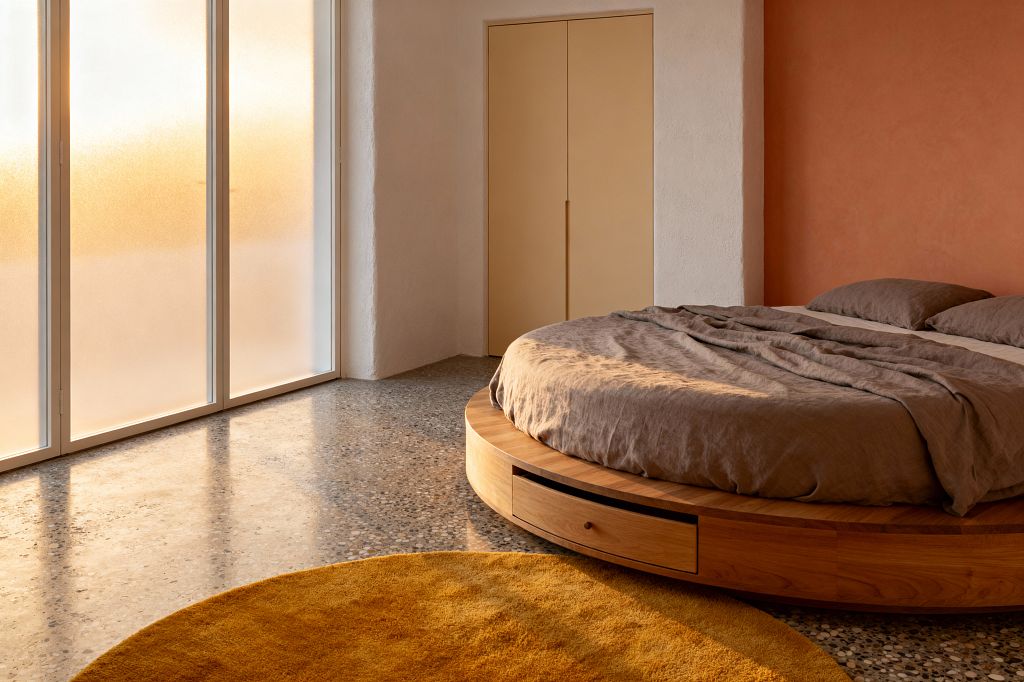 A minimalist kids' room features a low round wooden bed with built-in drawers and taupe linen bedding. The space has terrazzo flooring, a mustard circular rug, and floor-to-ceiling frosted glass doors that diffuse warm light. Walls are warm white with a terracotta accent section, and a flush beige closet sits in an alcove, creating a calm, clutter-free atmosphere focused on simple forms and soft textures.