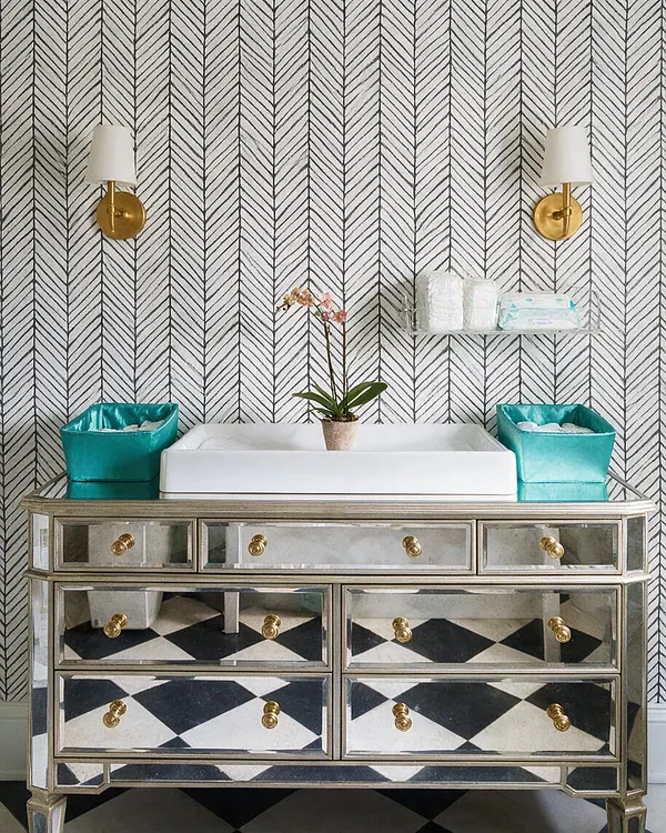 A glamorous nursery changing area with a mirrored dresser topped by a white changing pad, two teal storage bins, and a small orchid centered on top. Chevron wallpaper covers the wall behind two brass wall sconces with white shades, creating a polished Hollywood Regency look.