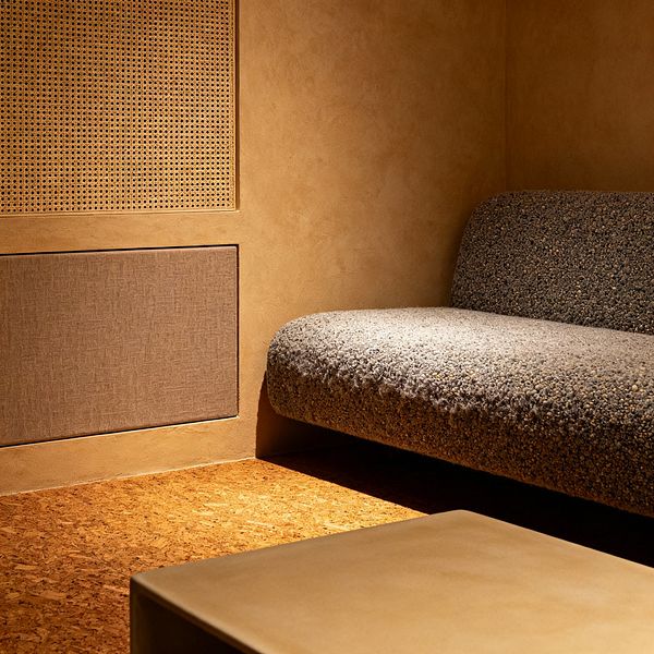 A compact minimalist media room with warm tan plaster walls and cork flooring features a low stone-gray boucle sofa with rounded edges and a simple low table. Two flush wall panels—a natural cane lattice and a taupe fabric insert—act as integrated acoustic treatments. Soft, indirect warm lighting creates a cocooned feel, emphasizing clean lines, earthy textures, and an uncluttered layout focused on comfort and sound quality.