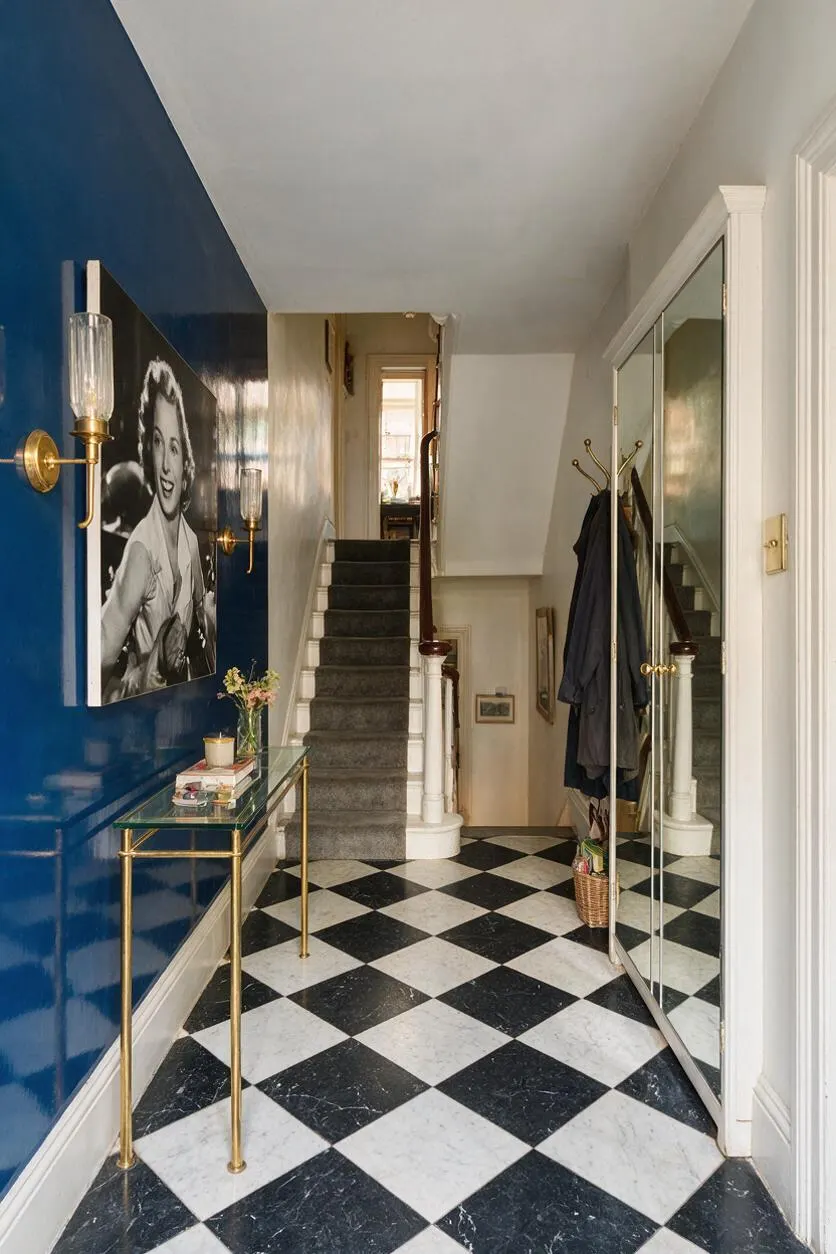 A Hollywood Regency entryway with a glossy cobalt blue accent wall, black-and-white checkered marble floor, and a large mirrored closet door. Brass wall sconces, a slim glass-top console, and a black-and-white portrait add glamour beside a carpeted staircase.