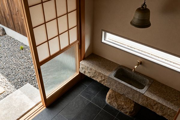 A minimalist Japanese-style laundry room features a rough-hewn granite counter with an inset stone sink and a simple brass wall-mounted faucet. A ceramic bell pendant hangs above; a long horizontal window and pale plaster walls bring soft light; dark slate floor tiles ground the space; and a wood-framed shoji sliding door with translucent washi and ribbed glass opens to a gravel garden and stepping stone.