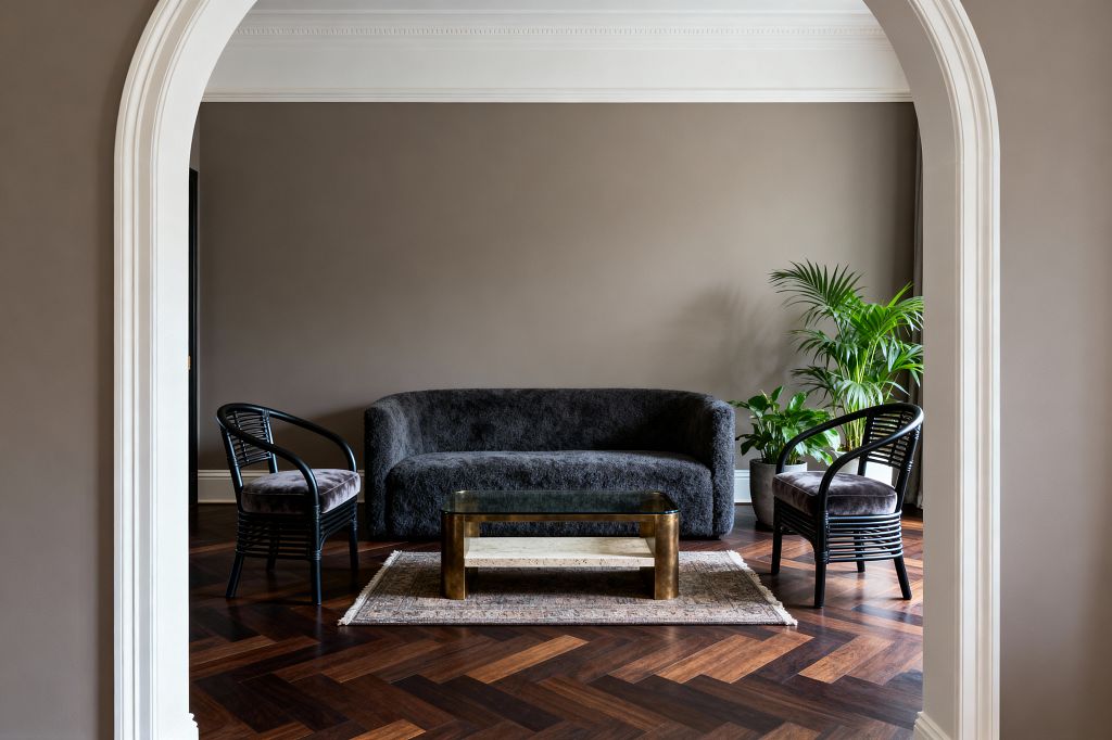 View through a white arched doorway into a calm living room with warm taupe walls and detailed crown molding. Centered is a curved charcoal bouclé sofa flanked by two black rattan armchairs with cushioned seats; a brass coffee table with a glass top and stone shelf sits on a small vintage rug atop walnut herringbone floors, with potted palms to the right and soft daylight filtering from an unseen window.