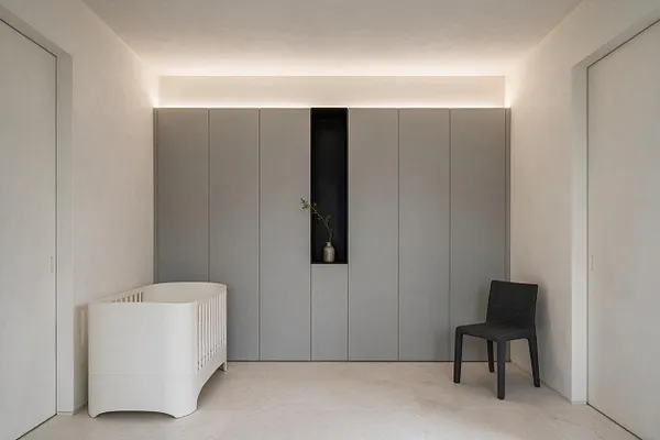 A minimalist nursery with a white crib on the left, a black chair on the right, and a wall of flat gray built-in cabinets with a narrow recessed niche holding a small vase. The room has white walls, a pale floor, and soft hidden lighting along the top of the cabinetry, creating a calm, uncluttered atmosphere.