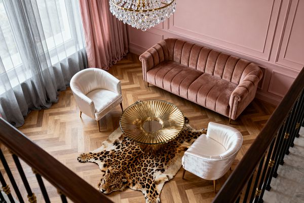 An overhead view of a Vintage Glam living room with blush pink paneled walls, a dusty-rose velvet channel-tufted sofa, and two ivory velvet barrel chairs around a fluted gold coffee table with a glass center. A leopard-print hide rug sits on oak herringbone floors beside tall windows with gray sheers and pink drapery, while a crystal chandelier sparkles above.