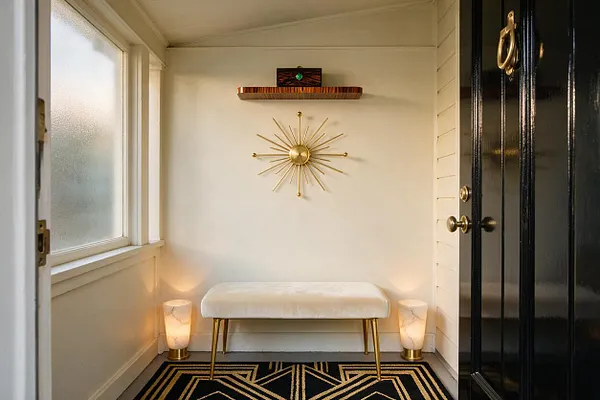 A small front porch styled in Art Deco fashion features a black glossy door with brass hardware, a cream upholstered bench with gold legs, and a black-and-gold geometric rug. A gold sunburst wall sculpture and two marble-like floor lamps create a symmetrical, elegant entryway with warm lighting.