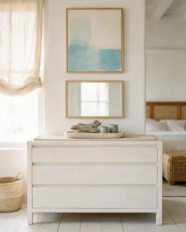 A serene coastal master bedroom corner with a light wood dresser topped by woven and ceramic decor, two framed artworks above it, and sheer curtains filtering daylight. In the background, a bed with a woven headboard and a rattan bench reinforce the relaxed, airy style.