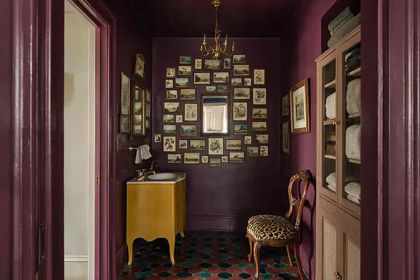 A compact powder room with deep plum walls and ceiling, a small yellow vanity with a round sink, and a brass chandelier overhead. The back wall is filled with a dense gallery of framed botanical and landscape prints, while a leopard-print chair and built-in cabinet with folded towels add layered, maximalist detail.