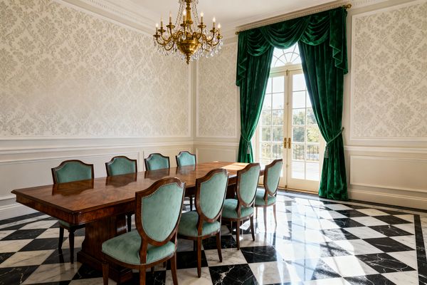 A formal dining room with a long polished mahogany table and eight mint‑green upholstered chairs with nailhead trim. The room features a black‑and‑white checkerboard marble floor, ivory damask wallpaper set within panel molding, and an ornate brass chandelier with crystal drops. Emerald velvet curtains frame tall French doors with brass hardware, filling the space with daylight and garden views.