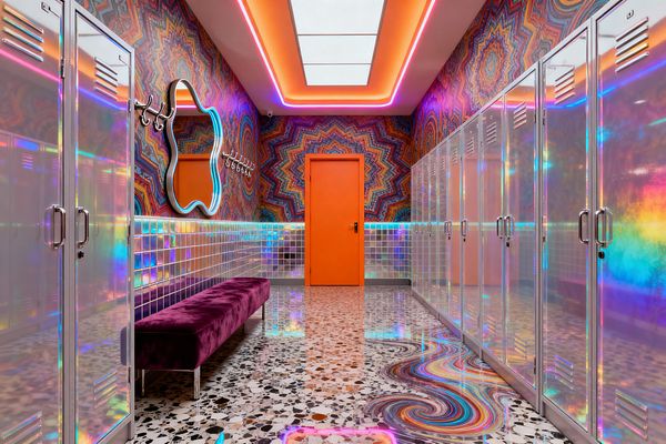 A vivid mudroom features rows of iridescent metal lockers flanking a terrazzo floor with a rainbow resin swirl. A purple velvet bench sits beneath a wavy LED-outlined mirror and prismatic glass-tile wainscot, while kaleidoscope starburst wallpaper and a bright orange door glow under pink-and-orange cove lighting around a central ceiling panel, with chrome coat hooks lining the wall.