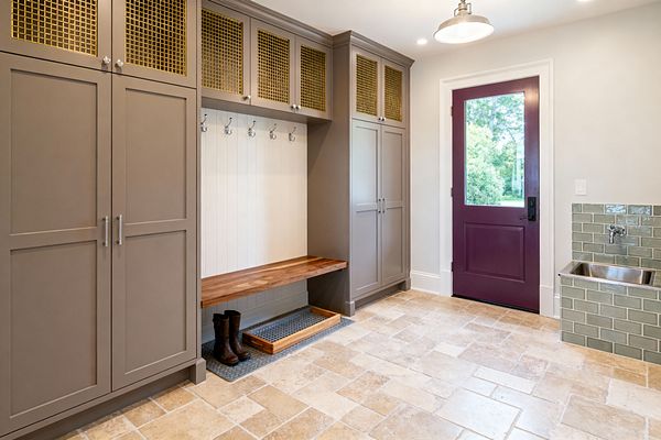 A transitional mudroom with greige Shaker cabinets featuring brass mesh uppers frames a white shiplap nook with metal hooks and a floating walnut bench over a boot tray. Tumbled travertine floors lead to a deep plum door with a glass panel, while a stainless utility sink is set into sage green subway tile, lit by a brushed metal pendant and recessed ceiling lights.