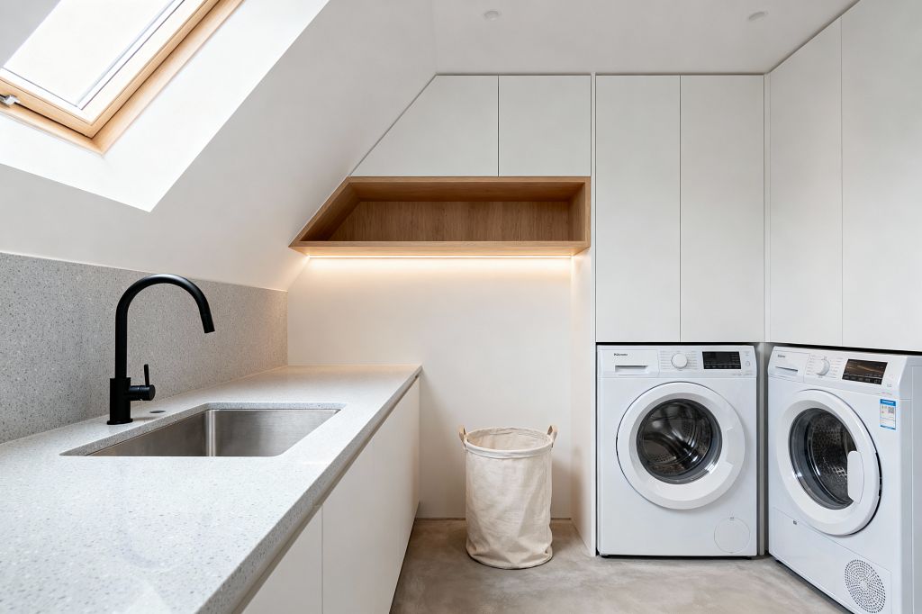 A minimalist laundry room set under a sloped ceiling features a large skylight, white handleless cabinets, and a light gray quartz worktop with matching upstand. A stainless-steel undermount sink is paired with a matte black gooseneck faucet. An oak-lined open shelf with warm LED strip lighting sits above a canvas laundry hamper. Side-by-side white front-loading washer and dryer stand next to tall cabinetry on a smooth gray concrete floor.
