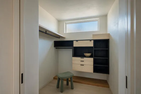 A compact closet in Japandi style with white walls, a high frosted window, and a floating storage system combining light oak fronts with matte black open shelving. A slim hanging rail runs along the left wall, and a small upholstered stool sits on the light floor, creating a calm, minimal dressing space.