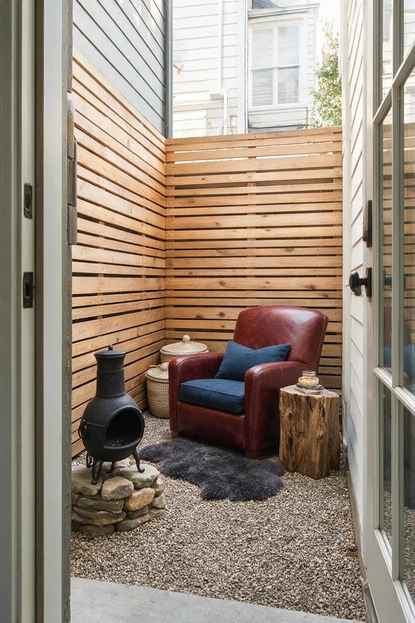 A small rustic backyard seating nook enclosed by horizontal wood slat fencing, with a burgundy leather armchair, blue cushion, black chiminea, woven baskets, and a tree stump side table. The ground is covered in gravel with a dark sheepskin-style rug, creating a warm, intimate outdoor retreat.