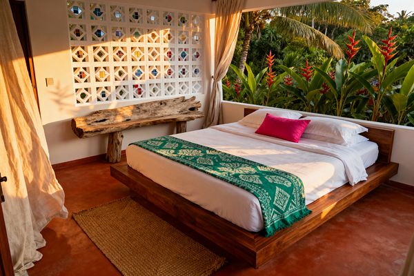 An open-air tropical bedroom shows a low wooden platform bed with white sheets, an emerald patterned throw at the foot, and a bright fuchsia lumbar pillow. Warm sunset light washes a terracotta-toned concrete floor, a jute rug, and a rustic driftwood bench; a white geometric breeze-block wall with small colored glass pieces and soft linen curtains opens to dense greenery with red heliconia flowers and palm trees.