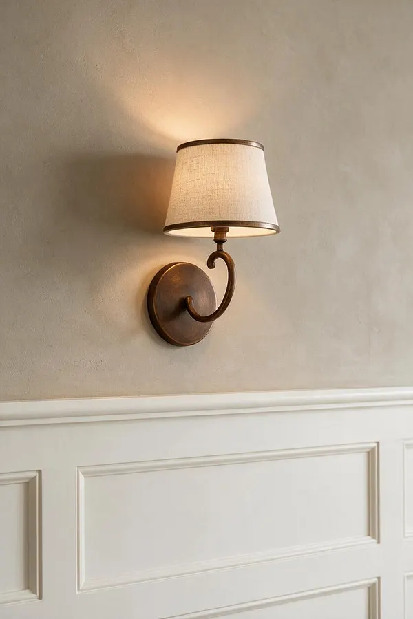 A transitional wall sconce with a curved bronze arm and round backplate is mounted on a warm beige wall above white wainscoting. The small tapered linen shade casts a soft glow, creating a classic and elegant accent.