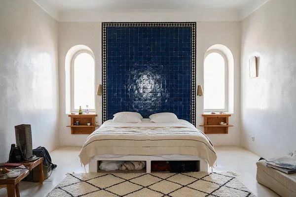 A guest bedroom with a low bed centered against a glossy deep blue tile feature wall, flanked by two arched windows and small carved wood shelves. The room has white plaster walls and floors, a cream striped blanket, a black-and-cream geometric rug, and simple floor cushions and woven accents that give it a Moroccan-inspired feel.