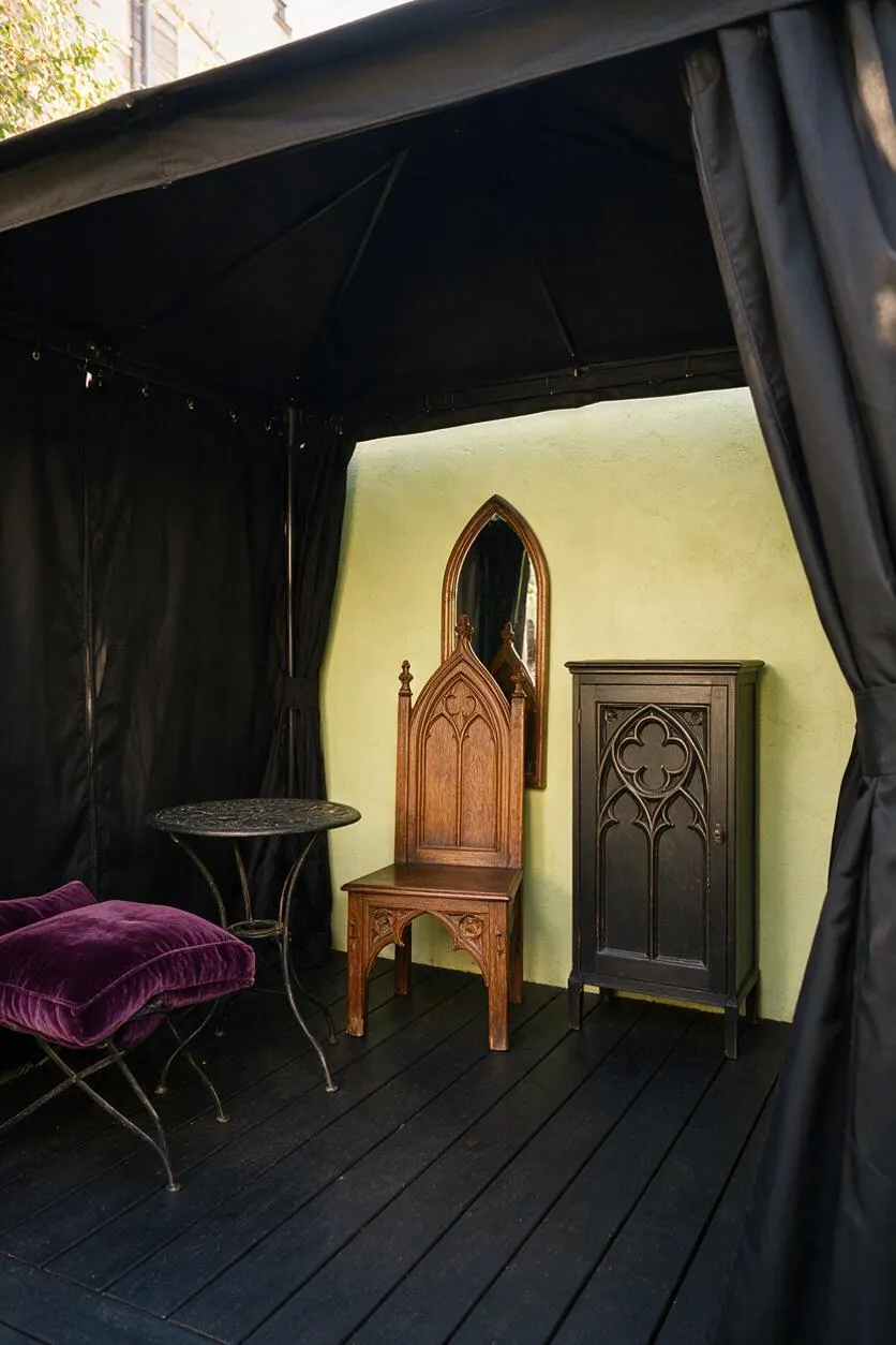 A small backyard seating area styled in Gothic decor, with black canopy drapes, a black painted floor, and a pale green wall as the backdrop. The space includes a carved wooden throne-style chair, a dark cabinet with Gothic arch detailing, a round metal side table, and a purple velvet cushion for rich contrast.