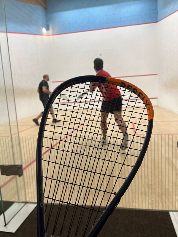A new squash racket is prominently displayed in the foreground while two players engage in a match in the background.