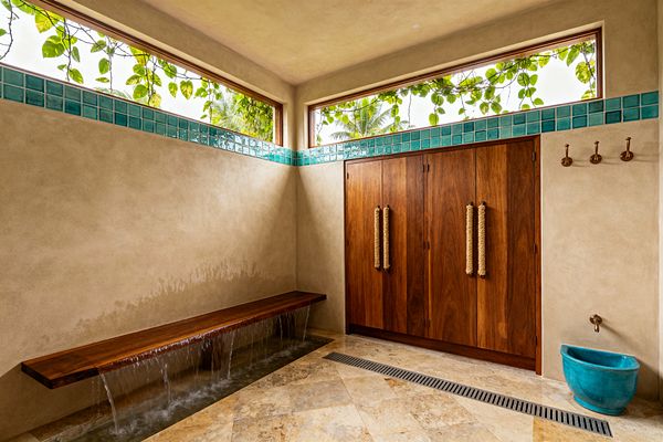 A light-filled tropical mudroom features sandy plaster walls, clerestory windows framed by leafy vines, and a narrow band of turquoise glass tiles. A floating teak bench spills water into a shallow rinse channel over diagonal travertine, opposite tall teak cabinets with oversized rope pulls, brass wall hooks, and a small bronze spigot above a teal ceramic bucket, creating an airy, resort-like entry space.