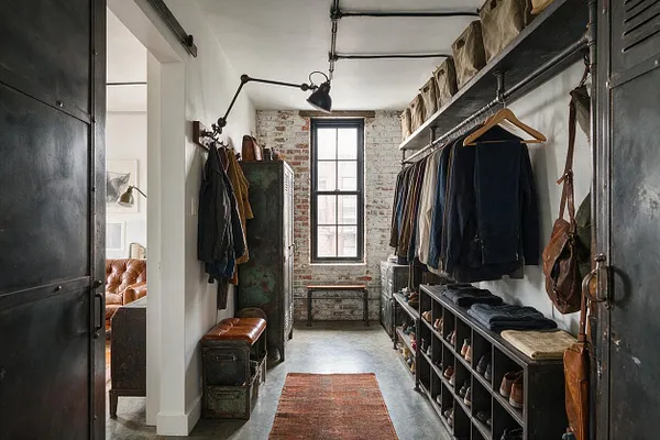 A narrow industrial-style closet with exposed whitewashed brick walls, black metal doors, and a concrete floor. The space includes open clothing racks, metal shelving with shoes and folded clothes, vintage trunks, a leather bench, and a tall window bringing in natural light.