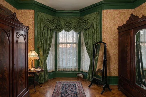 A Victorian-style closet room with dark carved wooden wardrobes on both sides, a tall freestanding mirror, and a bay window dressed in layered green curtains and lace sheers. The room has patterned wallpaper, deep green trim, a small side table with a lamp, a narrow oriental rug, and a polished wood floor.