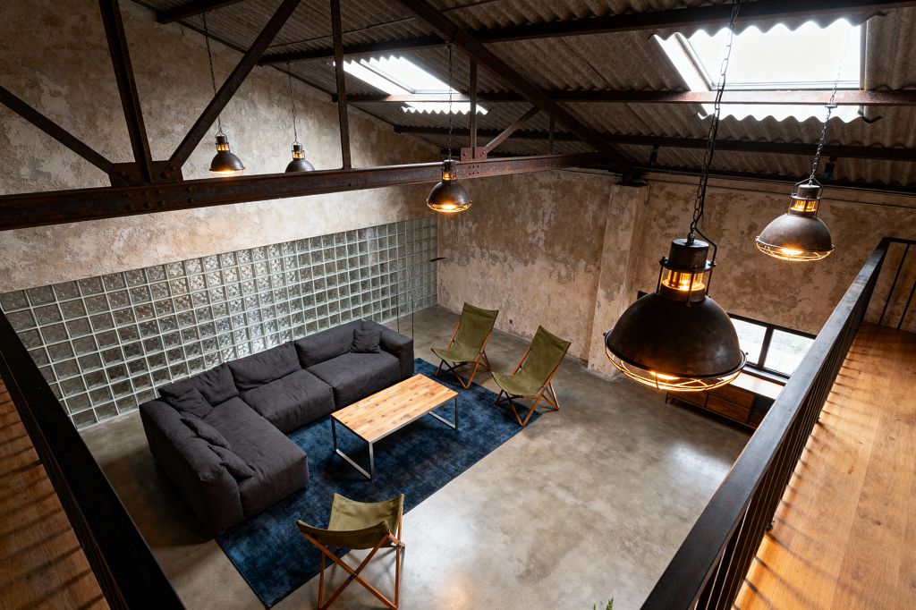 A double-height industrial living room viewed from a mezzanine shows raw plaster walls, a polished concrete floor, and exposed steel trusses under a corrugated roof with skylights. A charcoal sectional sits on a deep blue rug with a wood-and-steel coffee table and three olive sling chairs, lit by large dome pendant lights; a full glass-block wall and a low window with a wood console bring in diffuse natural light.