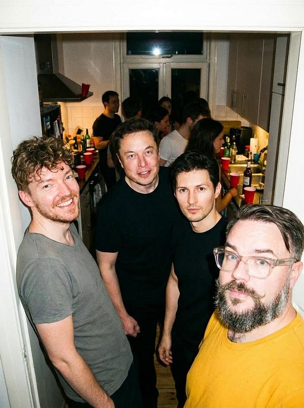 A group of four men poses in a kitchen filled with people and party supplies.