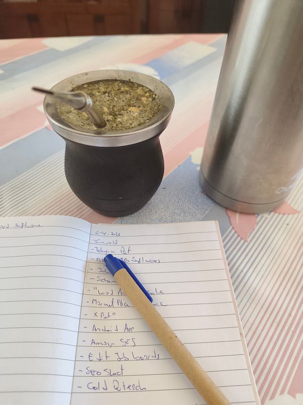 A flat-laid workspace image featuring a cup of yerba mate with a bombilla, a laptop, a notepad with handwritten tasks, and a pen on a wooden surface.