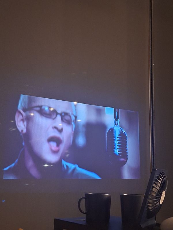 A projection of a singer performing, with two mugs and a fan visible in the foreground.