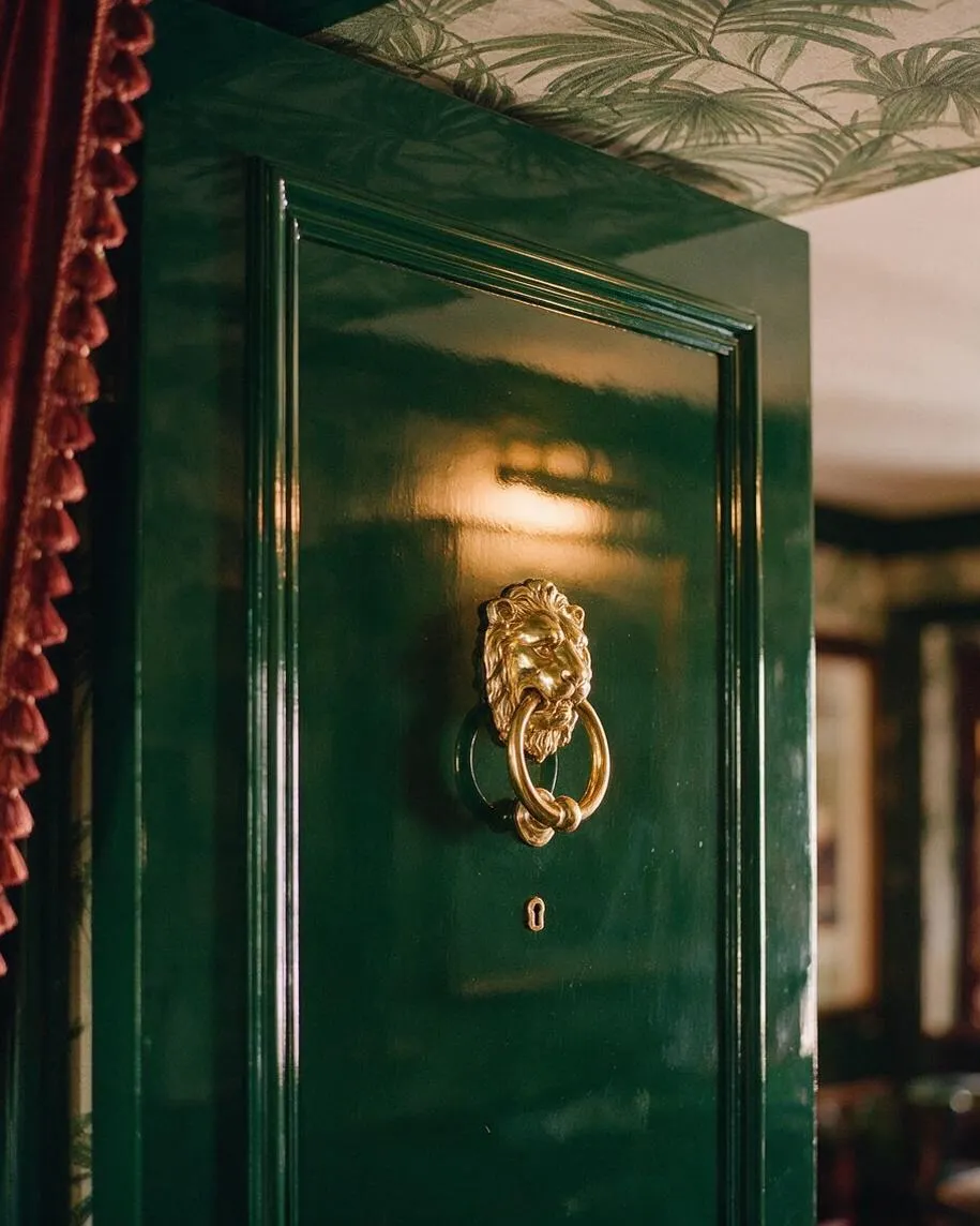 A close-up of a glossy dark green paneled door with a brass lion head knocker and ring handle. The surrounding space includes burgundy fringe detail on the left and a leafy patterned ceiling or wallpaper above, creating a rich maximalist look.