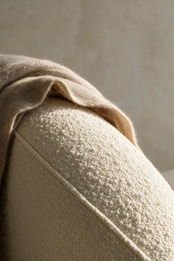 A close-up of a cream bouclé upholstered sofa arm draped with a soft camel-colored wool throw. The background is a warm neutral wall, creating a calm modern interior with tactile textures and understated tones.